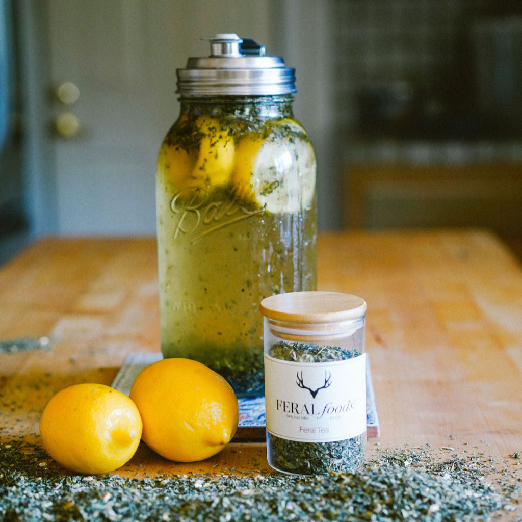 A mason jar filled with tea leaves and a infuser flask with tea leaves around, lemons to the side, all placed on a wooden surface.