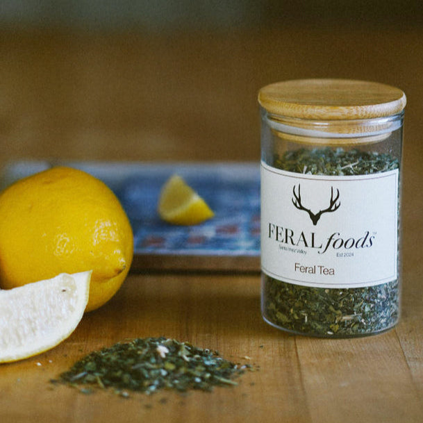 A jar of Feral Tea Blend with a wooden lid, placed on a wooden surface, accompanied by a slice of lemon and a whole lemon in the background, along with some scattered tea leaves.