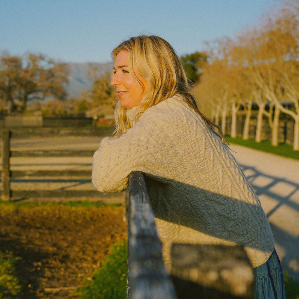 Woman leaning on a wooden fence in a scenic outdoor setting with trees and clear sky.