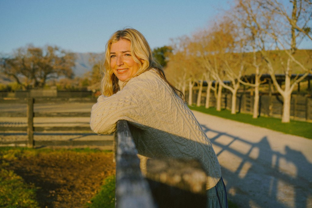 Keri Joy in a cream sweater leaning on a fence at golden hour