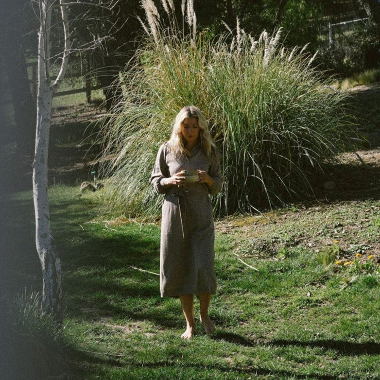 Woman in a long dress standing in a grassy area with tall plants and trees in the background.