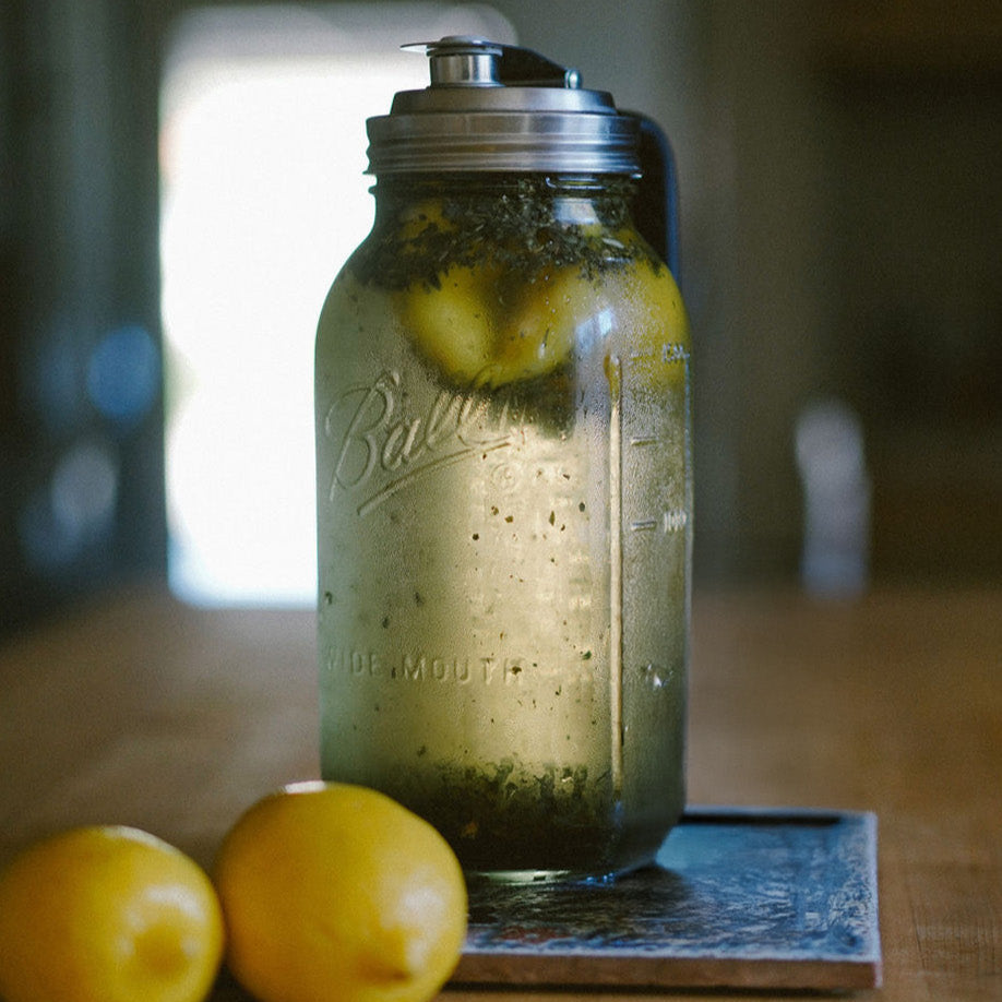 A clear glass mason jar filled with tea leaves and a slice of lemon on a wooden surface, with two lemons beside it.