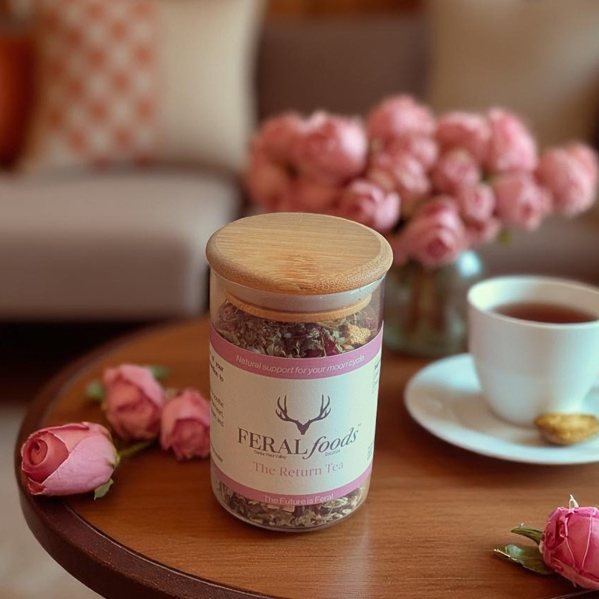 Jar of Feral Foods on a table with pink roses and a cup of tea in a cozy living room setting.